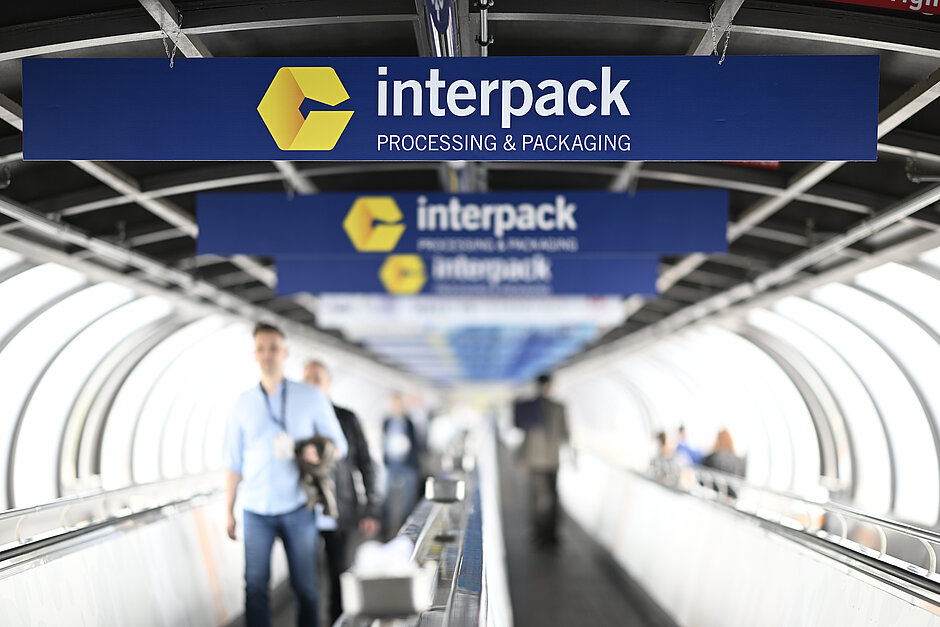 Des personnes marchant sur un trottoir roulant sous plusieurs panneaux bleus arborant le logo jaune d'interpack et la mention « interpack PROCESSING & PACKAGING », dans une structure en forme de tunnel.