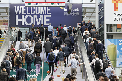 Une foule nombreuse emprunte les escaliers roulants en direction d'une pancarte sur laquelle on peut lire « BIENVENUE CHEZ VOUS » à l'entrée du salon Interpack.