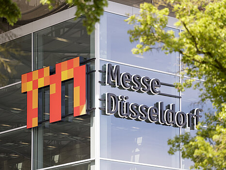 L'extérieur d'un bâtiment moderne en verre, avec le logo « Messe Düsseldorf » bien en évidence sur la façade, partiellement encadré par des branches d'arbres vertes.