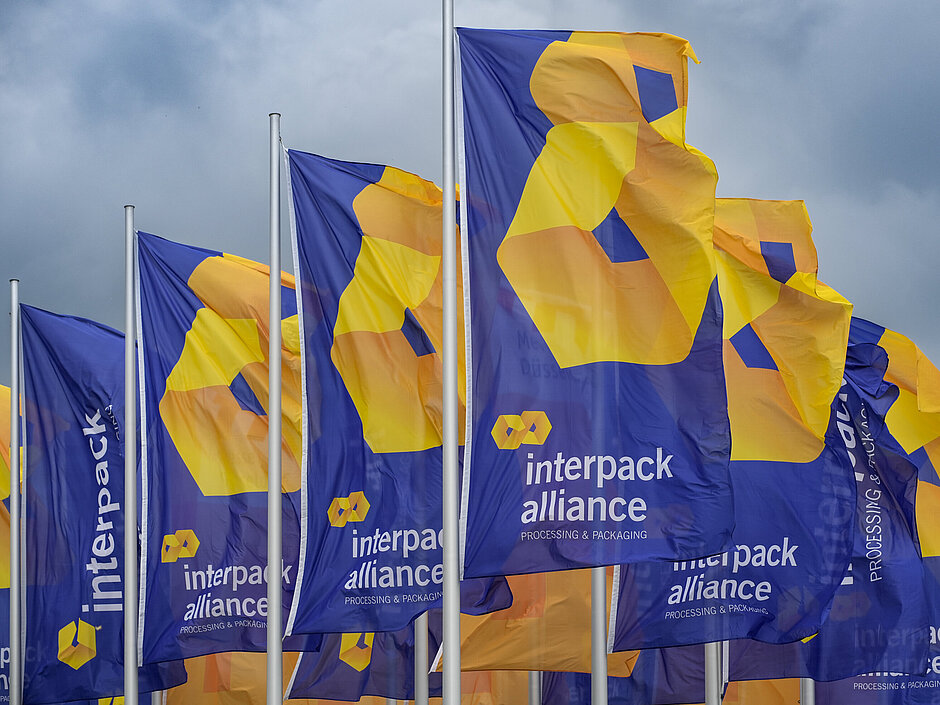 Plusieurs drapeaux bleus flottent au vent, arborant le logo jaune-orange et l'inscription « interpack alliance – Processing & Packaging » sur fond de ciel nuageux.
